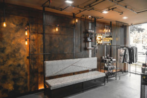 Industrial themed Barber shop waiting area. Rust painted wall with mesh and pipework with feature lighting. Pipework along ceiling and down the wall. Pipework used to create shelving and product display.